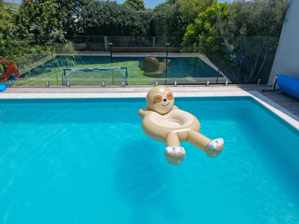 Pool Fence Compliance A bright backyard swimming pool with clear blue water, featuring an inflatable sloth float. A modern frameless glass pool safety fence with stainless steel spigots is in the foreground, ensuring compliance and style. A Pool Fence Inspection is ready to confirm this well-maintained barrier meets all safety standards.